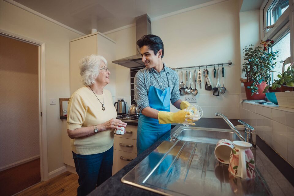 Teenage boy is talking to his grandmother while he washes the dishes in her kitchen.