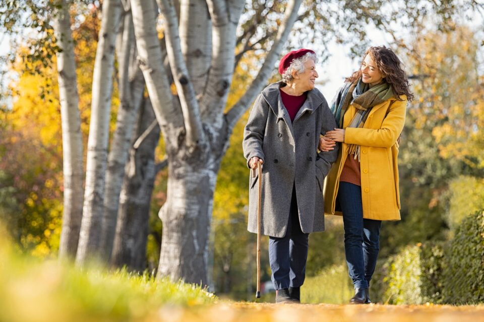 Granddaughter walking with senior woman in park wearing winter clothing. Old grandmother with walking cane walking with lovely caregiver girl in sunny day. Happy woman and smiling grandma walking in autumn park.